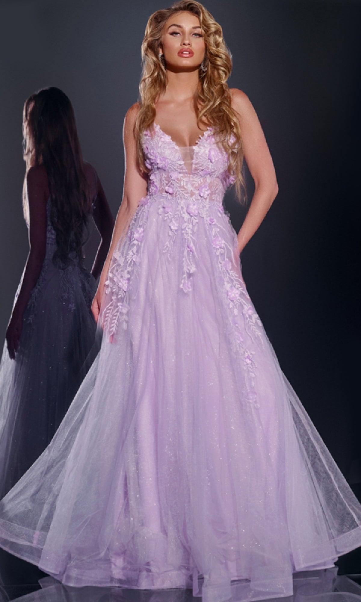 A woman stands before a mirror in the Jovani 40472 long prom dress, featuring lilac fabric with flowing lines, 3D floral accents, and lace embroidery that highlight both her look and its beautiful reflection.